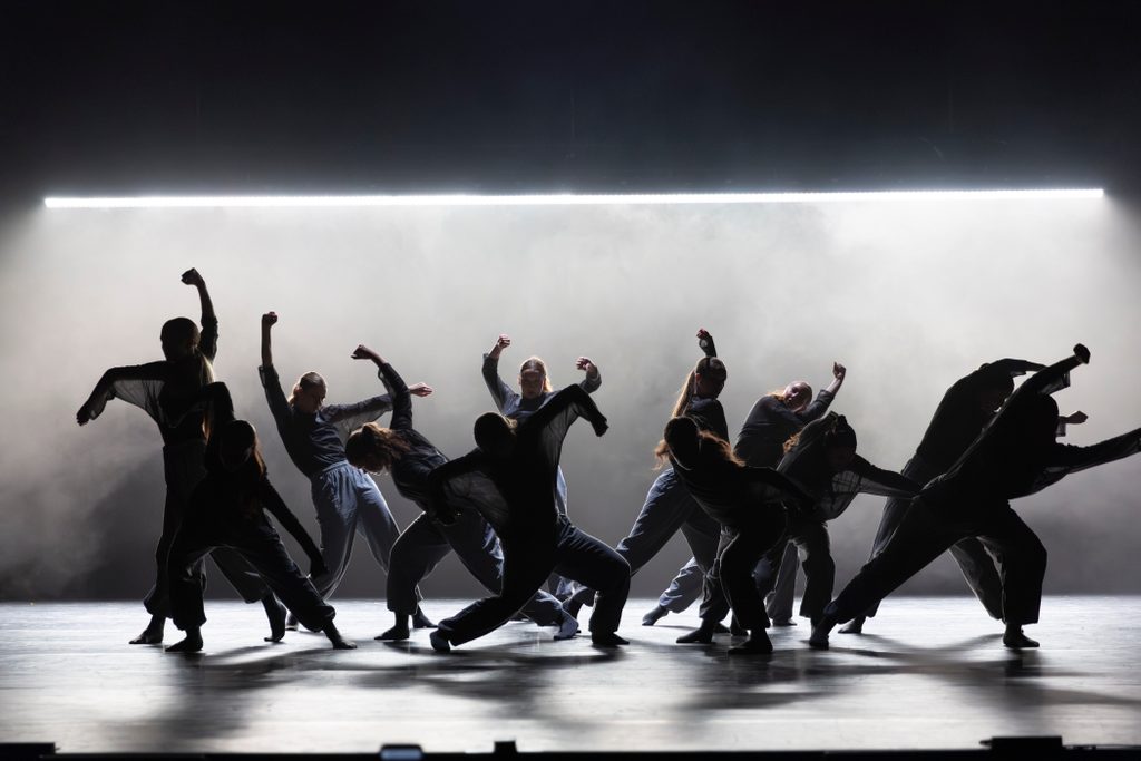 A group of dancers in dark clothing perform a synchronized routine on stage, silhouetted against a bright horizontal light and smoky background.