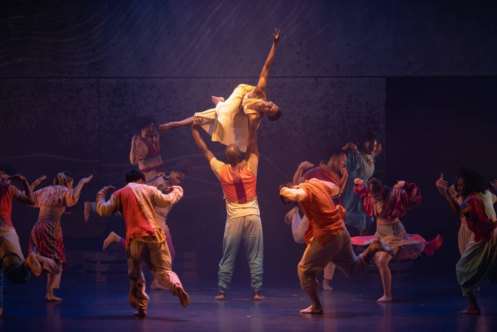 A group of dancers perform on stage, with one dancer lifting another high above their head while others dance energetically around them.
