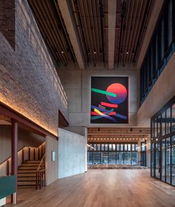Spacious modern interior with wooden flooring, high ceiling, and a large colorful abstract tapestry on the wall. Stairs and floor-to-ceiling windows are visible.