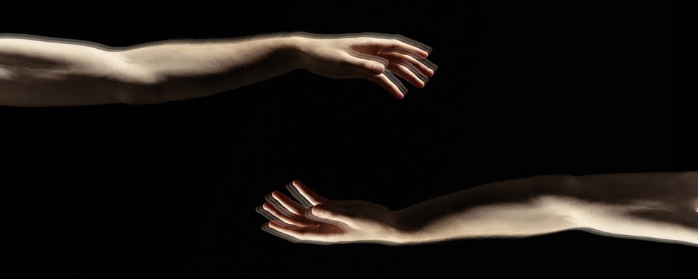 A black background with two arms reaching towards the centre. They have pale skin and relaxed hands.