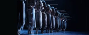 A group of female ballet dancers standing en pointe in a line on dark-lit stage. They wear long chiffon dresses.
