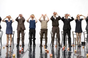 A group of jugglers in a big white room stand in front of chairs and hold up apples to their eyes