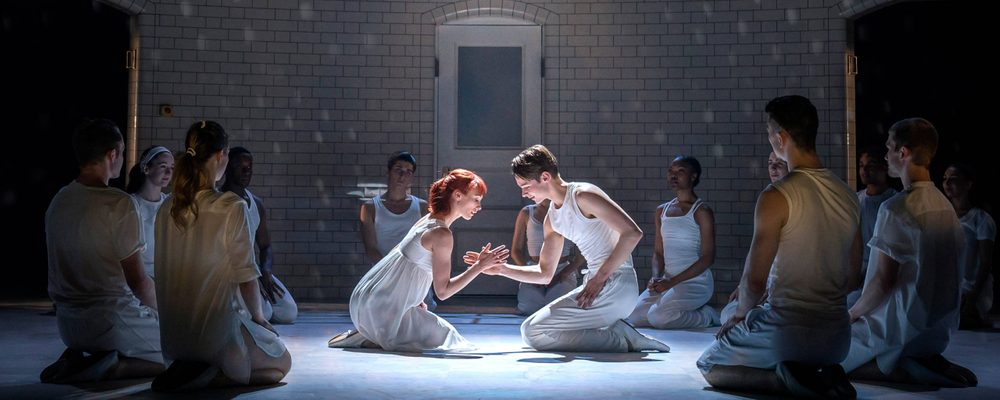 A room full of young adults sitting in a circle and in the middle holding hand sits two star crossed lovers