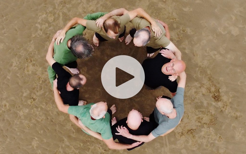 A group of 8 men on a beach stood in a circle with their hands around each other
