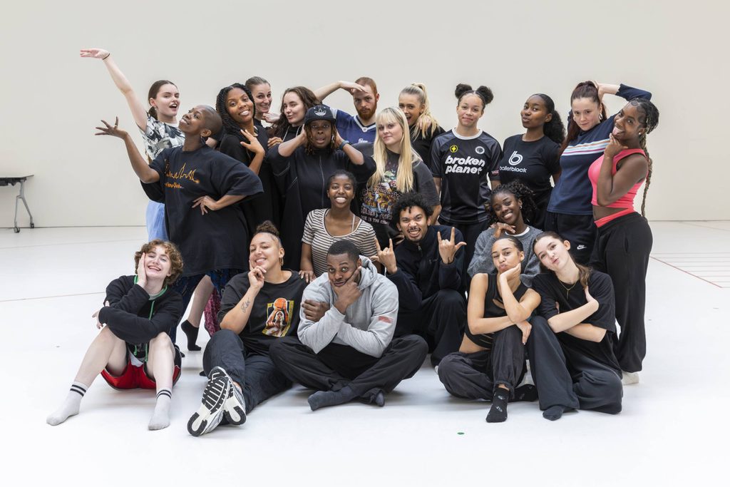 A group of young adults pose together in a dance studio, some standing and some sitting, making playful gestures and smiling at the camera.