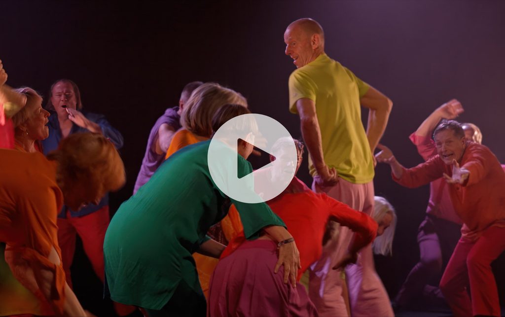 A large group of dancers all wearing colourful clothes dance together on stage
