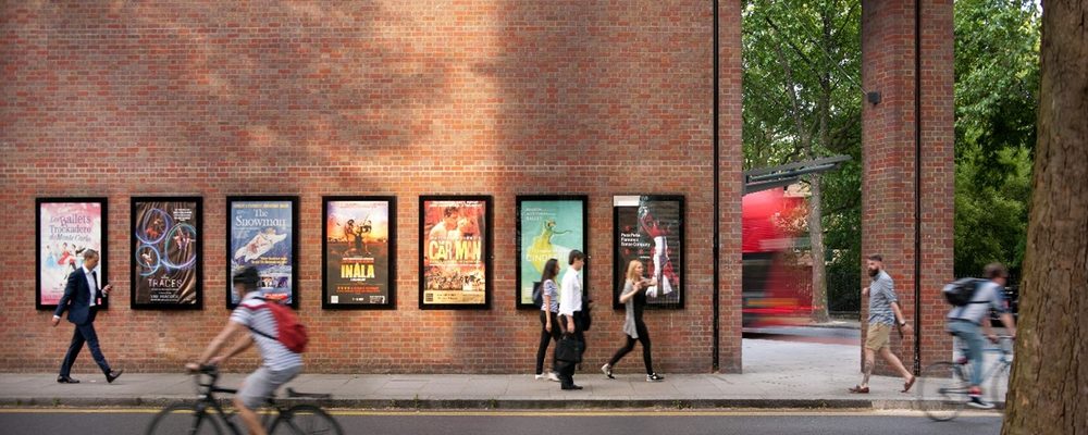 A photo of the outside wall of Sadler's Wells Theatre building with show posters and pedestrians and a cyclist passing by
