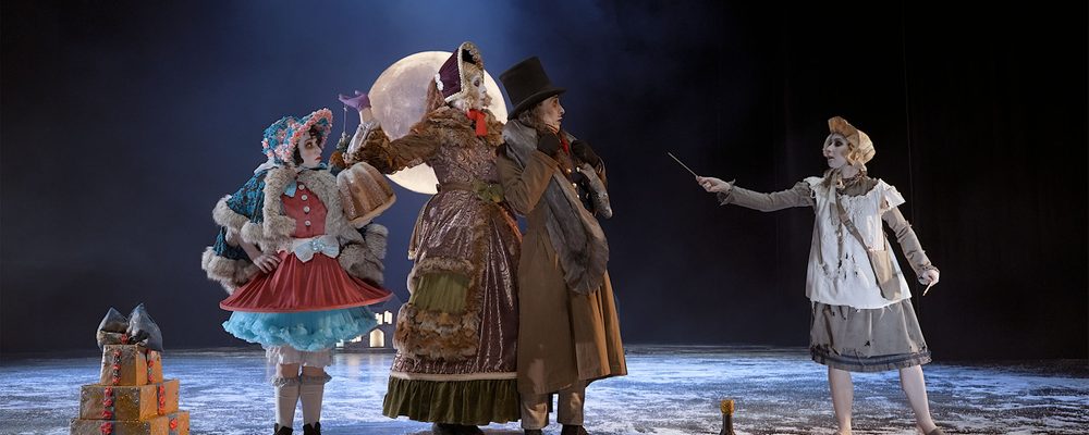 On a snowy winter night, three dancers, the Donnarumma family stand together looking to the right at the little match girl who is wearing a white bonnet and pinafore and is holding out a match.