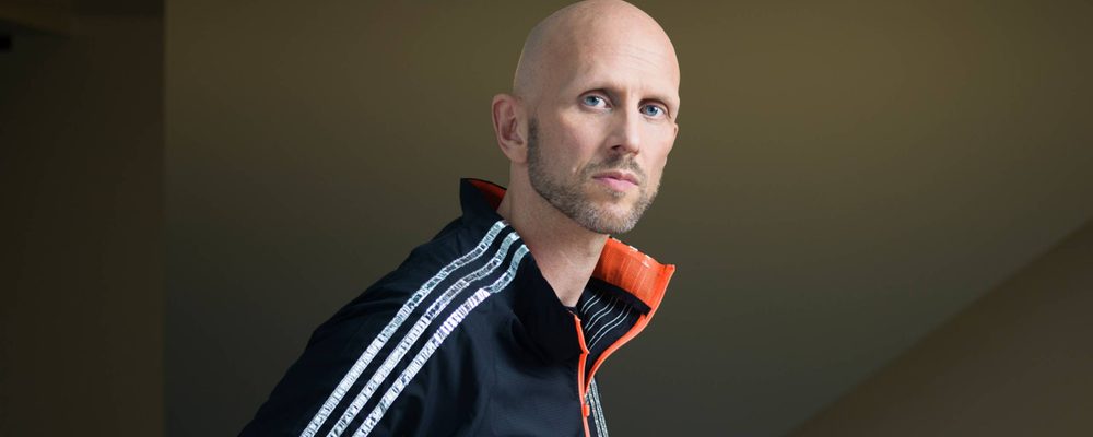 Wayne McGregor wears a sports top which is black with white stripes on the arm and orange inside the collar. He is a bald man and looks towards the camera.