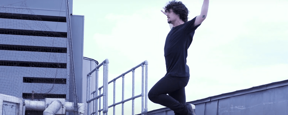 A photograph of Jesús Carmona performing a flamenco move, with his left up arm and his right arm to the side. He has dark curly hair and is wearing a black t-shirt.